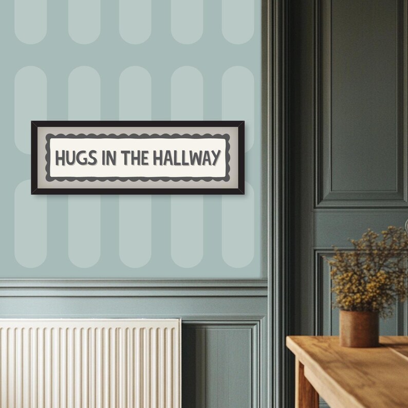 May include: A rectangular framed sign with the text "HUGS IN THE HALLWAY" in black letters. The sign has a scalloped border and is set against a light background. The frame is black. The sign is hanging on a light blue wall.