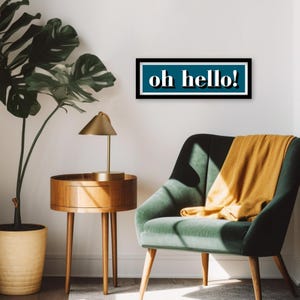 May include: A teal blue rectangular sign with a black frame and white text that reads "oh hello!" hanging on a white wall above a green velvet armchair with a yellow throw blanket.