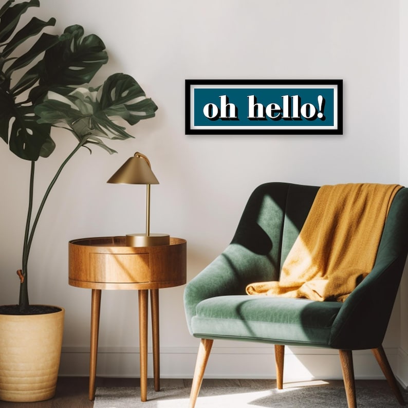 May include: A teal blue rectangular sign with a black frame and white text that reads "oh hello!" hanging on a white wall above a green velvet armchair with a yellow throw blanket.