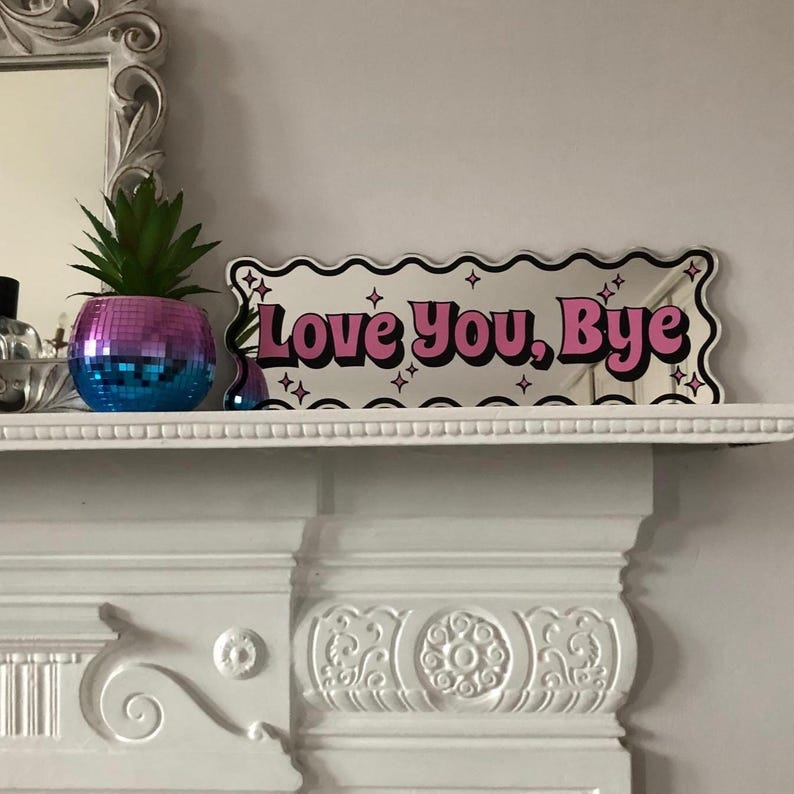 以下が含まれることがあります： ピンクと黒で「Love You, Bye」の文字が入った装飾ミラー。 ミラーは、星のアクセントが付いた波状の白いボーダーが付いています。 ミラーの左側には、小さなディスコボールスタイルのプランターと人工多肉植物があります。