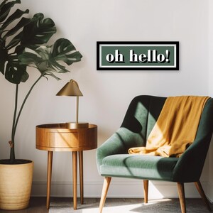 May include: A green and black framed wall art print with the text "oh hello!" in white letters. The print is hanging on a white wall above a green armchair with a yellow throw blanket.
