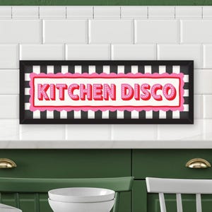 May include: A rectangular wall sign in a black frame featuring the text KITCHEN DISCO in pink, dotted letters. The sign has a white background with a black and white vertical striped border and a scalloped pink frame surrounding the text.