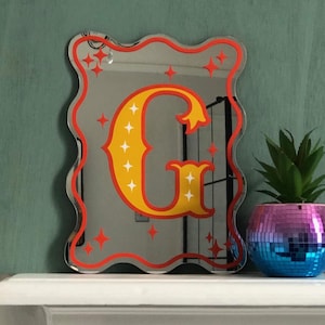 May include: A decorative mirror in a wavy shape with a red border and a large yellow letter "G" with white star accents. The mirror has red star accents around the edges. A small plant in a disco ball pot is to the right.