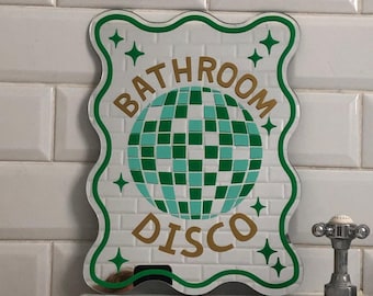 May include: A decorative sign with a white background and a green wavy border. The sign features the words "BATHROOM DISCO" in gold lettering, along with a disco ball graphic in shades of green and teal. Green star accents are also included.