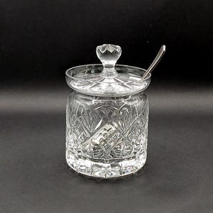 May include: A clear, cut-glass jar with a lid and a small spoon. The jar has a cylindrical shape with intricate leaf and diamond patterns. The lid features a decorative knob. The spoon rests on the lid.