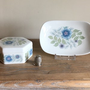 May include: A white ceramic trinket box and rectangular dish, both with a floral pattern in blue, green, and purple. A silver thimble is next to the box. The items are on a wooden surface.