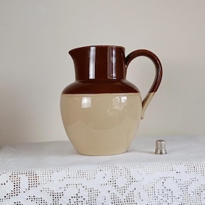 May include: A two-tone ceramic jug with a brown top and handle, and a beige body. A small silver thimble sits nearby. The jug has a classic design, suitable for serving beverages or as a decorative item. The jug is approximately 25 cm tall.
