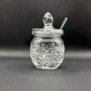May include: A clear, cut-glass sugar bowl with a lid and a small spoon. The bowl has a diamond pattern and a pointed finial on the lid. The spoon is made of silver-coloured metal.