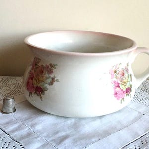 May include: A white ceramic bowl with a pink rim and handle, adorned with floral rose patterns in pink, yellow, and green. A silver thimble rests on a white lace doily. The bowl is likely vintage.