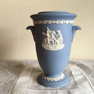 May include: A blue ceramic vase with white relief detailing. The vase features a classical scene with figures and a laurel wreath design. It has two handles and a pedestal base, suitable for home decor.