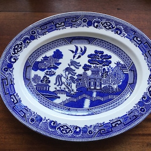 May include: Oval serving platter with a blue and white Willow pattern. The ceramic dish features a detailed landscape scene with pagodas, bridges, and trees. The rim has a decorative border. A classic piece for serving food.
