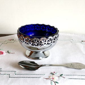 May include: A silver salt cellar with a cobalt blue glass insert and a decorative openwork rim. A small silver spoon rests beside the salt cellar. The items are on a white embroidered cloth.