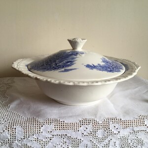May include: White ceramic serving bowl with a blue landscape design on the lid. The bowl has a scalloped edge and a decorative handle. The bowl is on a white lace tablecloth. The bowl is approximately 25 cm in diameter.