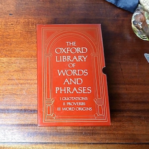 May include: A red book titled "The Oxford Library of Words and Phrases" with gold lettering. The cover features an arched design and lists sections on quotations, proverbs, and word origins. The book is on a wooden surface.