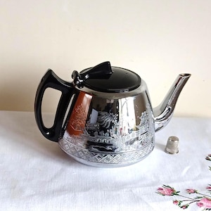 May include: A chrome teapot with a black handle and lid. The teapot has an engraved Asian-inspired design. A small silver thimble is next to the teapot. The teapot is likely made of metal.
