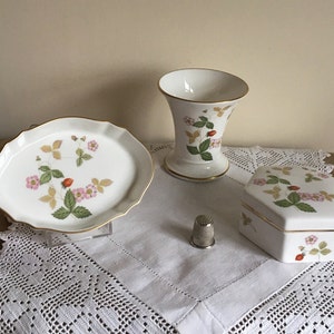 May include: A white ceramic plate, vase, and hexagonal box with a strawberry design. The plate has a scalloped edge and gold trim. The vase and box also have gold trim. A silver thimble sits on a white lace tablecloth.
