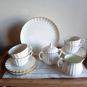 May include: A white porcelain tea set with gold edging, comprising cups, saucers, a large plate, a sugar bowl, and a creamer. The set is displayed on a wooden tray, ideal for serving tea.