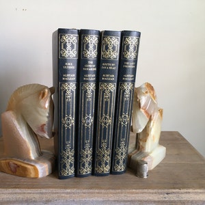 May include: Four hardback books with dark blue covers and gold lettering, including titles like "H.M.S. Ulysses" and "The Guns of Navarone." The books are held between two decorative onyx horse head bookends. A silver thimble rests on the wooden surface.