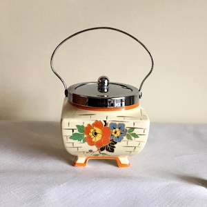 May include: A vintage ceramic sugar bowl with a silver lid and handle. The off-white body features a brick pattern and is decorated with orange, yellow, and blue floral designs. The base and rim have orange accents.
