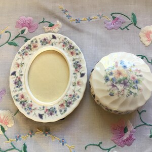 May include: An oval ceramic picture frame and a round trinket box, both adorned with floral patterns in pastel shades. The frame has a cream interior, and the box features a ridged design.