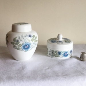 May include: Two white ceramic containers with blue floral patterns. One is a lidded jar, the other a small round box. A silver thimble sits nearby. The containers have a vintage aesthetic.