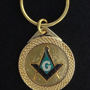 May include: Gold-colored keychain with a round, textured charm featuring a black and white Masonic symbol with a blue and white 'G' in the center.