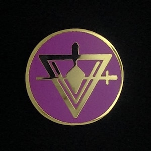 Könnte beinhalten: Eine goldfarbene Anstecknadel mit einem violetten Kreis und einem schwarzen und goldenen geometrischen Design im Inneren. Das Design zeigt ein Dreieck mit einer Diamantenform in der Mitte.