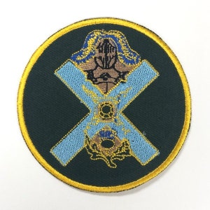 May include: A round, green embroidered patch with a gold border. The patch features a blue and white cross with a gold knight in the center. The knight is wearing a helmet and holding a sword. The cross is surrounded by a gold thistle.