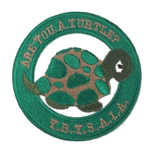 May include: Green embroidered patch with a cartoon turtle in the center. The turtle is brown and green with a white eye. The text around the turtle reads "Are you a turtle? Y.B.Y.S.A.L.A."