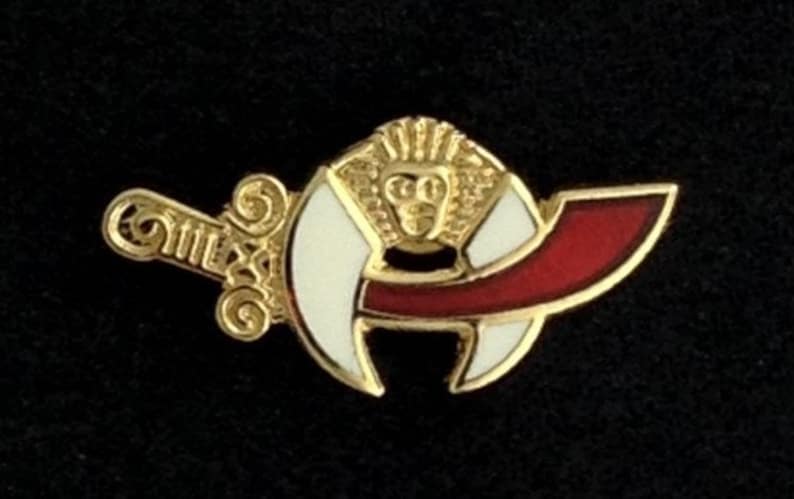 Puede incluir: Pin de color dorado con una luna creciente blanca y una cinta roja. El pin presenta una imagen estilizada de una cabeza con una corona.