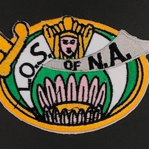 May include: A white, green and yellow embroidered patch featuring a woman wearing a crown and a banner that reads "Sons of NA".