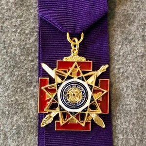 Peut inclure: Ruban violet avec fermoir doré et médaille décorative. La médaille présente un motif d'éclat d'étoile rouge et or, un emblème central et des épées croisées.