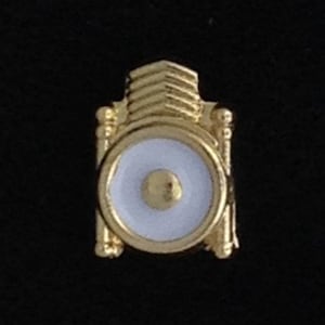 Könnte beinhalten: Eine goldfarbene Anstecknadel mit einem weißen Emaille-Zentrum und einem goldfarbenen Punkt in der Mitte. Die Anstecknadel ist glockenförmig.