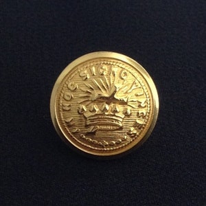 Pode incluir: Um botão dourado com uma coroa e um design de sol estilizado. O texto "SIGNO" está inscrito ao redor da borda do botão.
