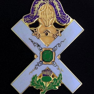 May include: A gold-colored pin with a white cross and a green gemstone in the center. The cross is surrounded by a gold chain and a purple and gold knight's helmet. A green thistle with a purple flower is at the bottom of the pin.