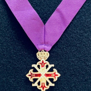 May include: A gold-colored cross-shaped pendant with red accents, suspended from a wide, purple ribbon. The pendant features a crown at the top and intricate detailing. The background is a dark, textured surface.