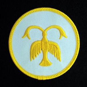 May include: A white circular patch with a yellow embroidered bird design. The bird has two outstretched wings and a long tail. The bird is surrounded by a yellow border.