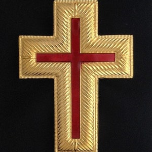 Może przedstawiać: Złoty krzyż z czerwonym środkiem. Krzyż ma fakturowaną powierzchnię i podniesioną krawędź.