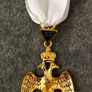 May include: A gold-colored Masonic medal with a white ribbon. The medal features a double-headed eagle with a crown, a triangle with "33°", and the Latin phrase "Deus Meumque Jus".