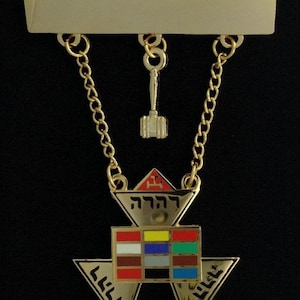 Könnte beinhalten: Goldener Freimaurer-Anhänger mit einem dreieckigen Design, das ein rot-weiß-blau-gelb-grün-braun-grau-schwarz kariertes Muster aufweist. Der Anhänger ist an einer goldenen Kette mit einer goldenen Stange und einem kleinen goldenen Hammer befestigt.