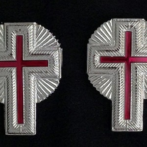 May include: Two silver-colored crosses with red enamel centers. The crosses are surrounded by a decorative, radial pattern.