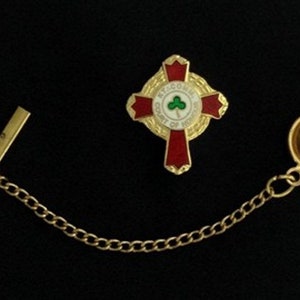 Op de afbeelding: Gouden stropdasgesp met een rood kruis en een witte cirkel met een groene klaver en de tekst "Welcome Court of Honor". De stropdasgesp is bevestigd aan een gouden ketting met een gouden sluiting.