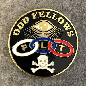 May include: A gold-colored metal pin with a black background. The pin features the words "ODD FELLOWS" in a gold-colored font, a stylized eye with rays of light, three interlocking rings with the letters "F", "L", and "T", and a skull and crossbones.