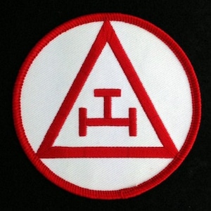 May include: A white embroidered patch with a red circle border. Inside the circle is a red triangle with a red symbol of a horizontal bar with two vertical bars on either side.