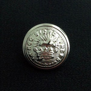 May include: A shiny, silver-colored button with a detailed embossed design. The button features a crown at the center, surrounded by intricate patterns and text. The button is circular and appears to be made of metal.