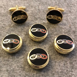 May include: Gold-toned cufflinks and shirt studs with a black face. The face of each accessory features the word "FLOT" in white, blue, and red. The cufflinks have a classic bar closure.