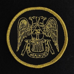 May include: A black embroidered patch with a gold border. The patch features two golden eagles with wings spread, a triangle with the number 32 inside, and a golden lyre with the text "MEA IN DEO EST".