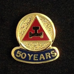 Könnte beinhalten: Goldene und blaue Anstecknadel mit einem roten Dreieck und dem Text "50 JAHRE". Das Dreieck enthält ein schwarzes Symbol.