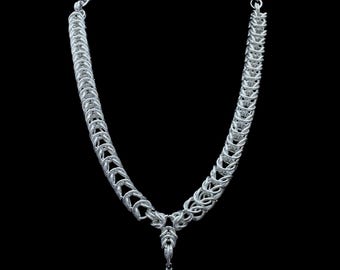 May include: A silver-toned necklace with a unique chainmail design. The necklace features a central pendant with a pointed, dagger-like charm. The chain is composed of interwoven metal rings, creating a textured appearance against a black background.