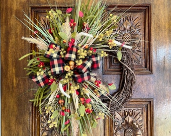 May include: A fall-themed wreath with a plaid bow, dried grasses, and red berries. The wreath is made of twigs and is hanging on a wooden door.